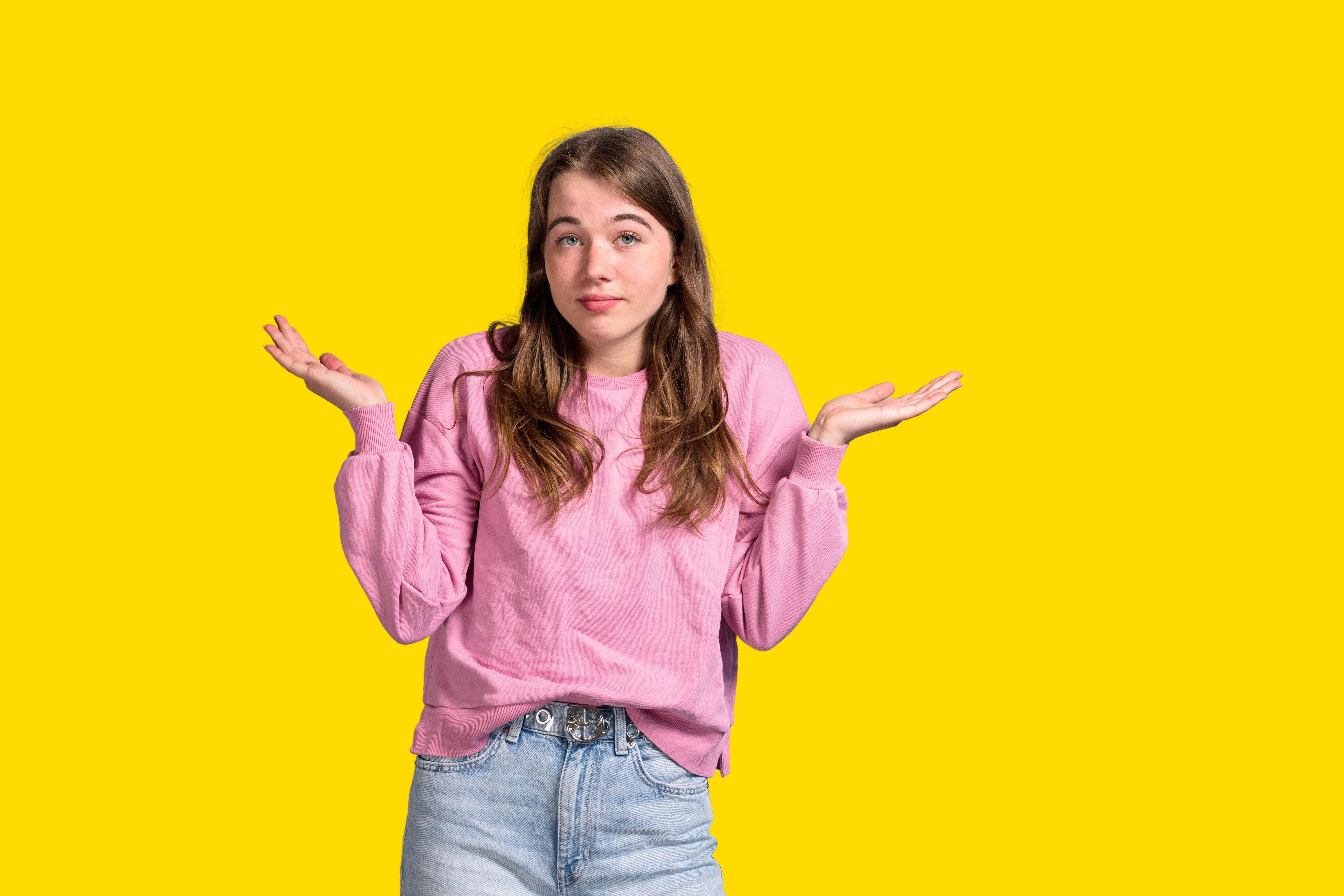 Student haalt haar schouders op, wat onzekerheid of twijfel uitdrukkend, tegen een effen gele achtergrond