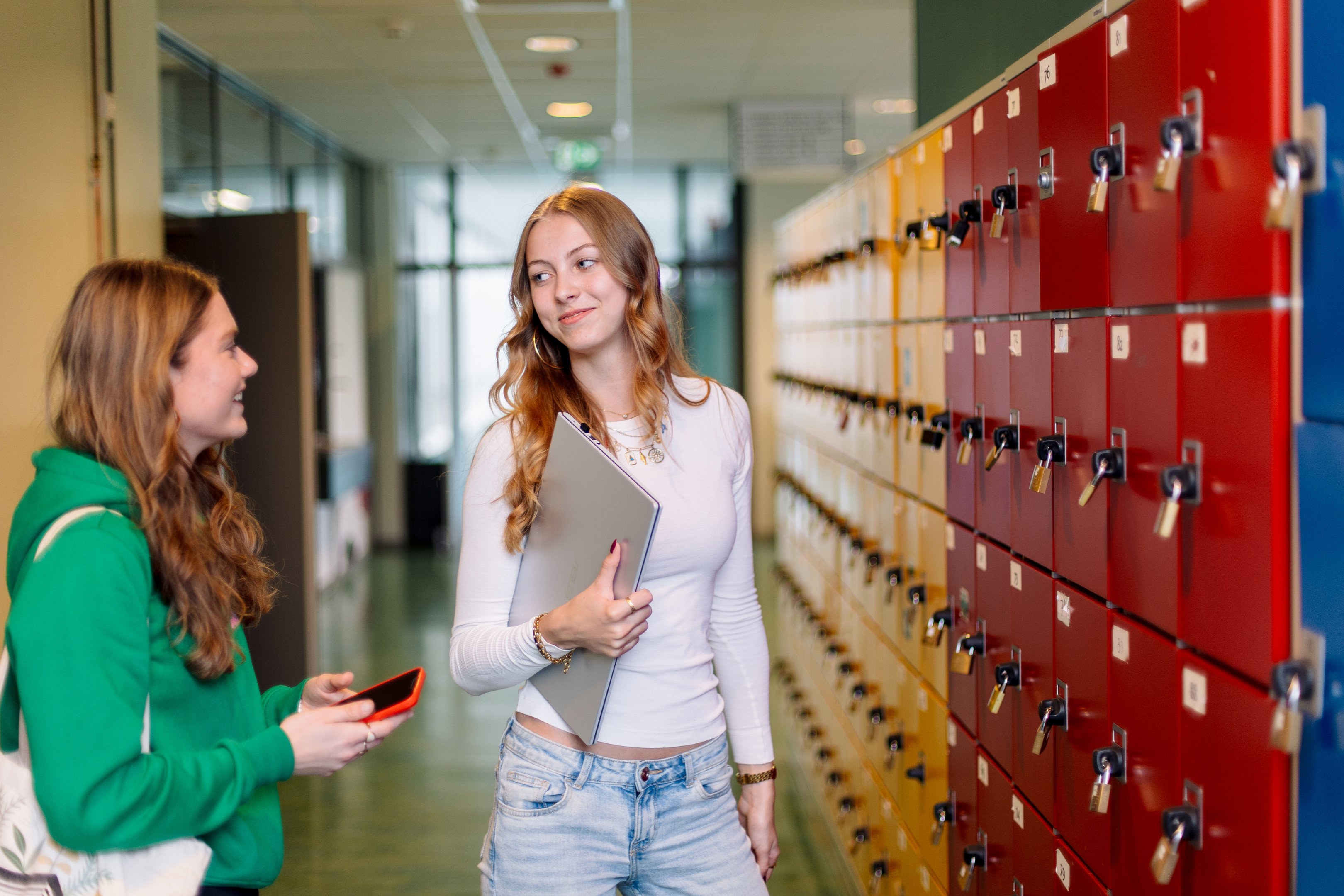 Twee studenten lopen langs kluisjes in een schoolgang, één met laptop en één met mobiel in gesprek