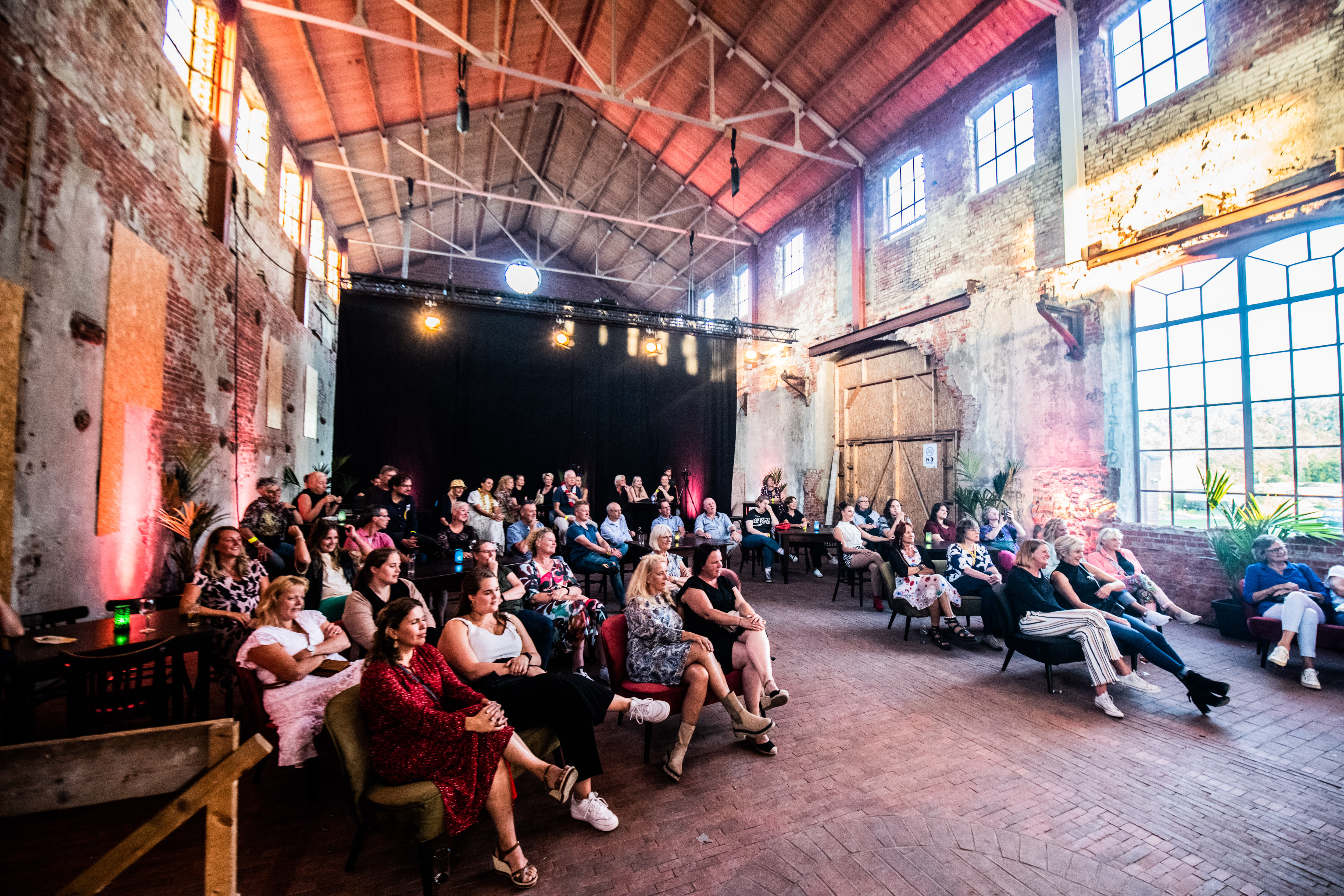 Groep bezoekers luistert aandachtig tijdens jubileumevenement Noorderpoort in grote industriële zaal.