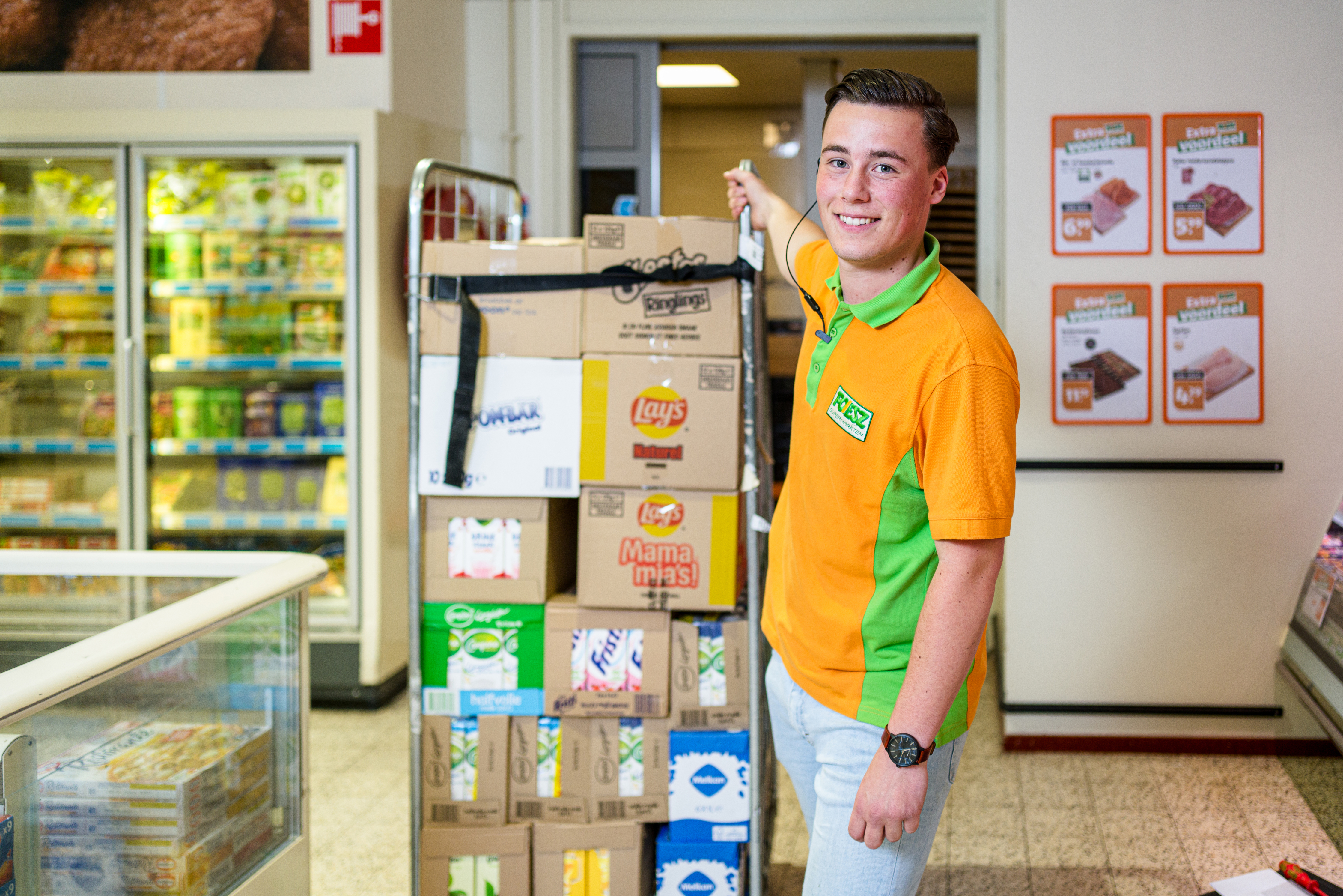Student helpt met voorraadbeheer in een supermarkt, stapelt dozen met diverse producten op een steekwagen.