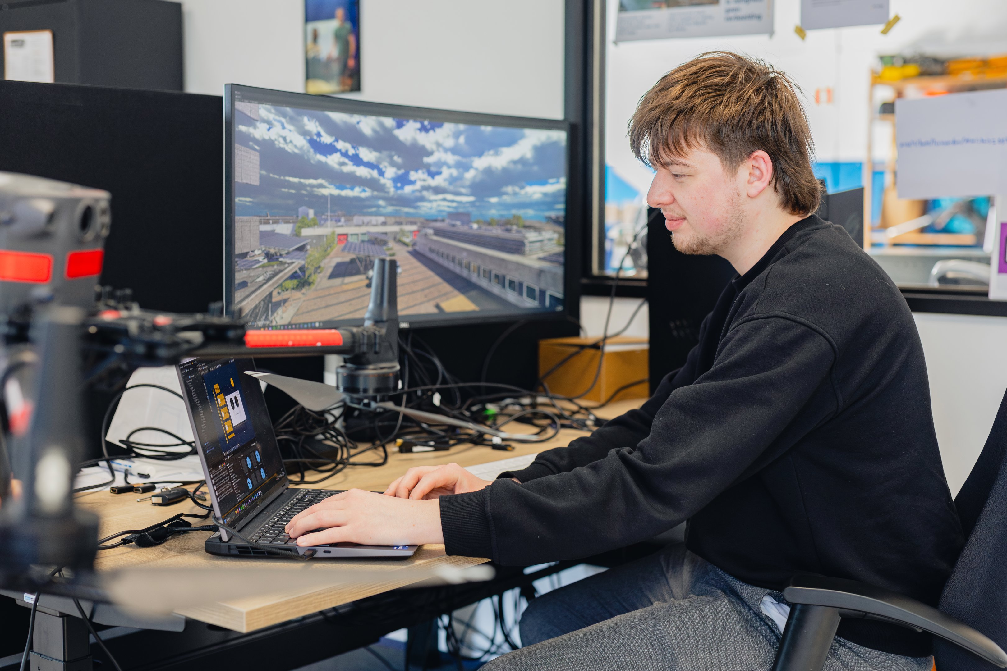 Student werkt met dronebesturingssoftware en bekijkt luchtbeelden van een industrieterrein op schermen.
