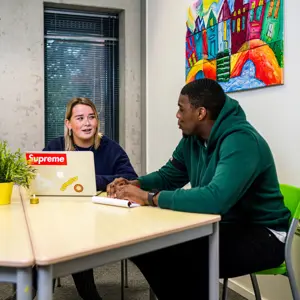 Twee personen voeren een gesprek aan een tafel, mogelijk over maatschappelijke zorg of begeleiding.