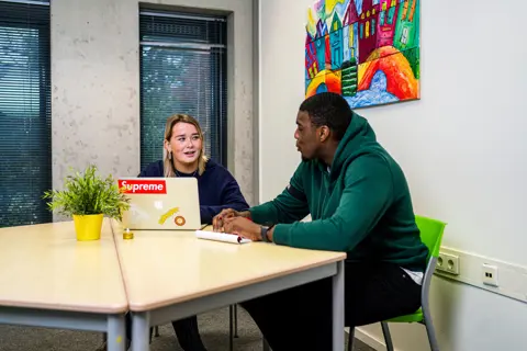 Twee personen voeren een gesprek aan een tafel, mogelijk over maatschappelijke zorg of begeleiding.