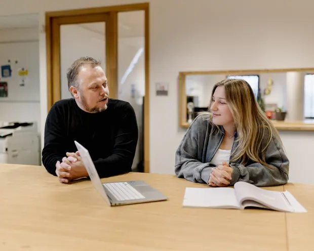 Twee volwassenen in gesprek aan tafel met laptop en open boek, gericht op competentie-erkent proces.
