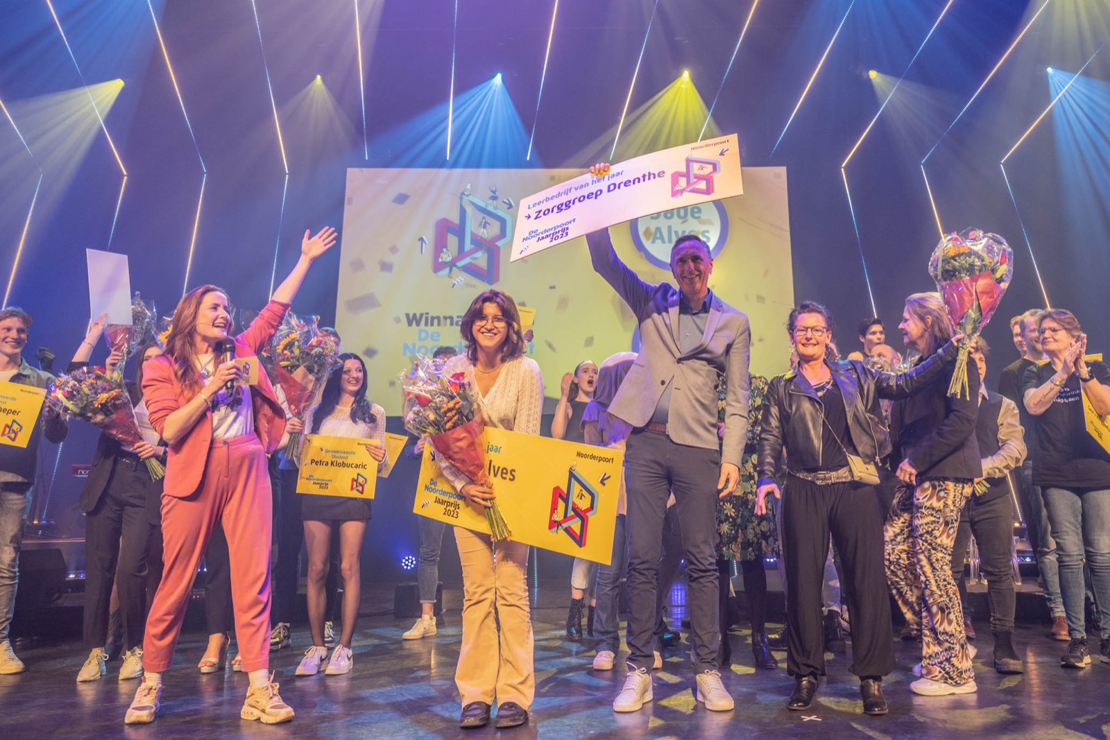 Studente Jade ontvangt de Noorderpoort Jaarprijs 2023 op het podium, omringd door feliciterende collega’s en medestudenten