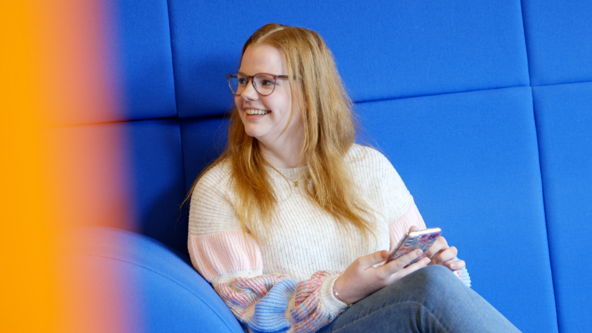 Student bekijkt mobiel en lacht terwijl ze zit in een leeromgeving met comfortabele zitplaats