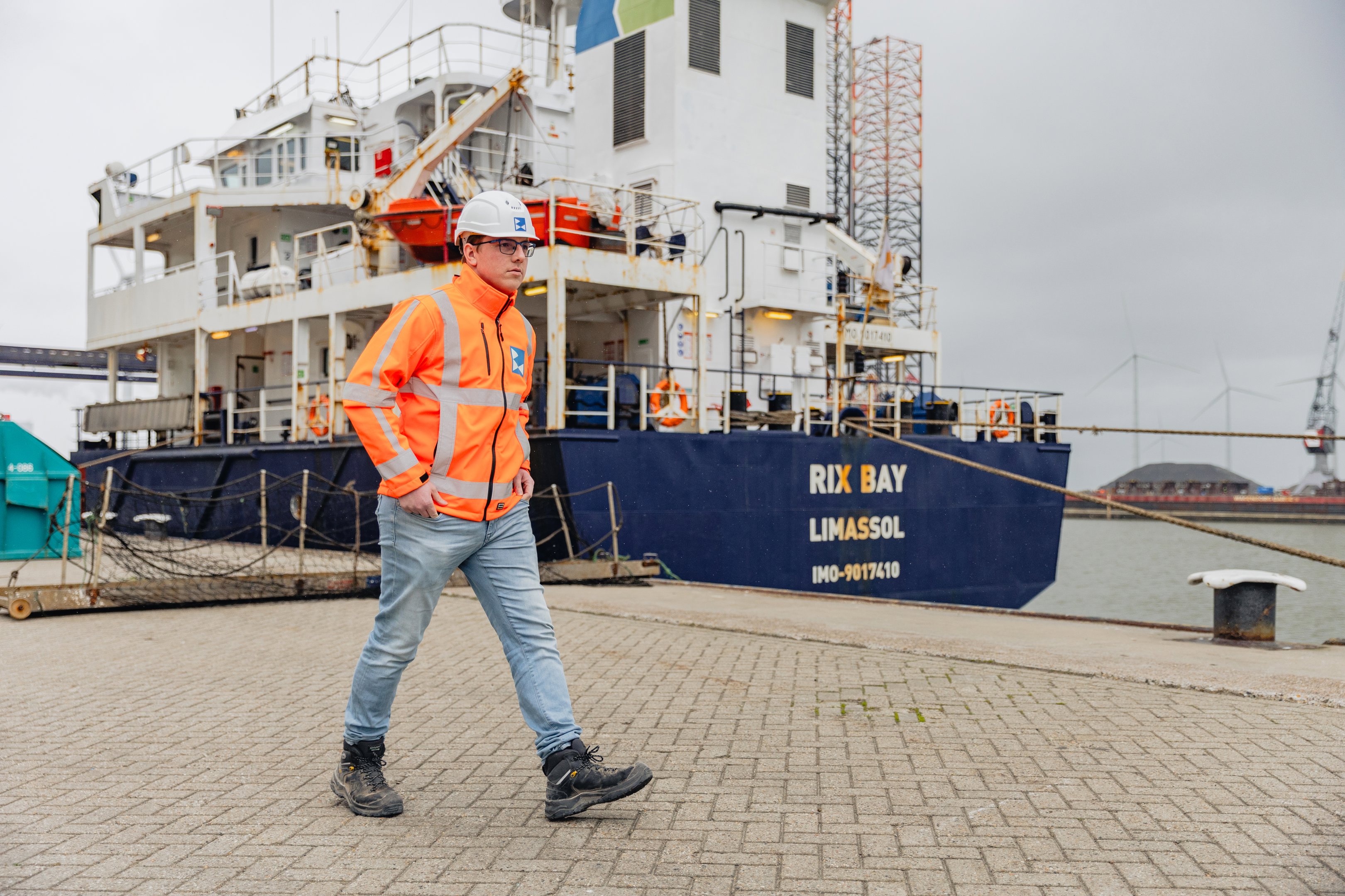 Student maritiem beroep loopt in werkjas en helm langs een schip in havengebied voor praktijkopleiding