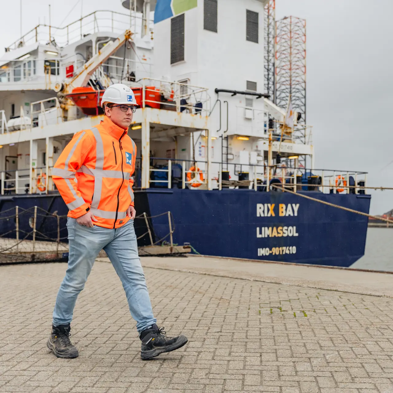 Student maritiem beroep loopt in werkjas en helm langs een schip in havengebied voor praktijkopleiding