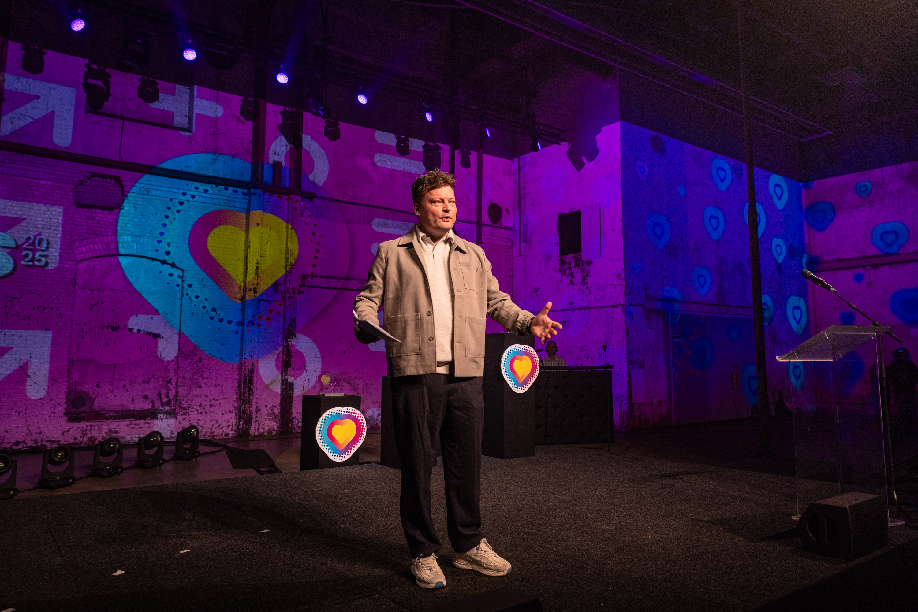 Presentator op podium tijdens Noorderpoort Jaarprijs 2025 met kleurrijke projecties van hartvormige symbolen op de achtergrond