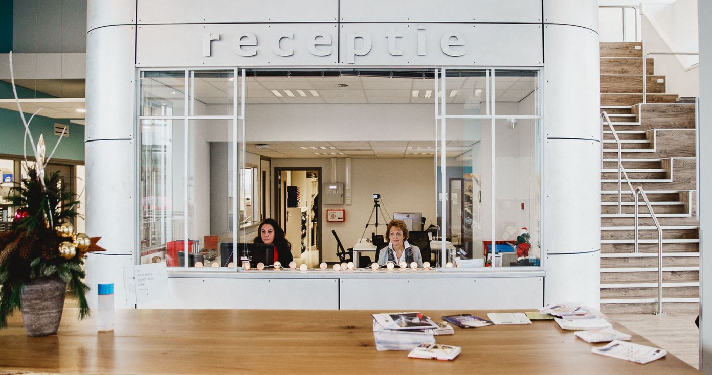 Balie van de receptie in het onderwijsgebouw aan Muntinglaan 3, met twee medewerkers achter het glas.