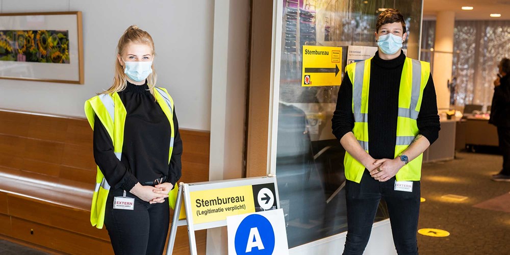 Twee studenten met veiligheidsvesten en mondkapjes begeleiden kiezers bij een stemlokaal tijdens verkiezingsdag.