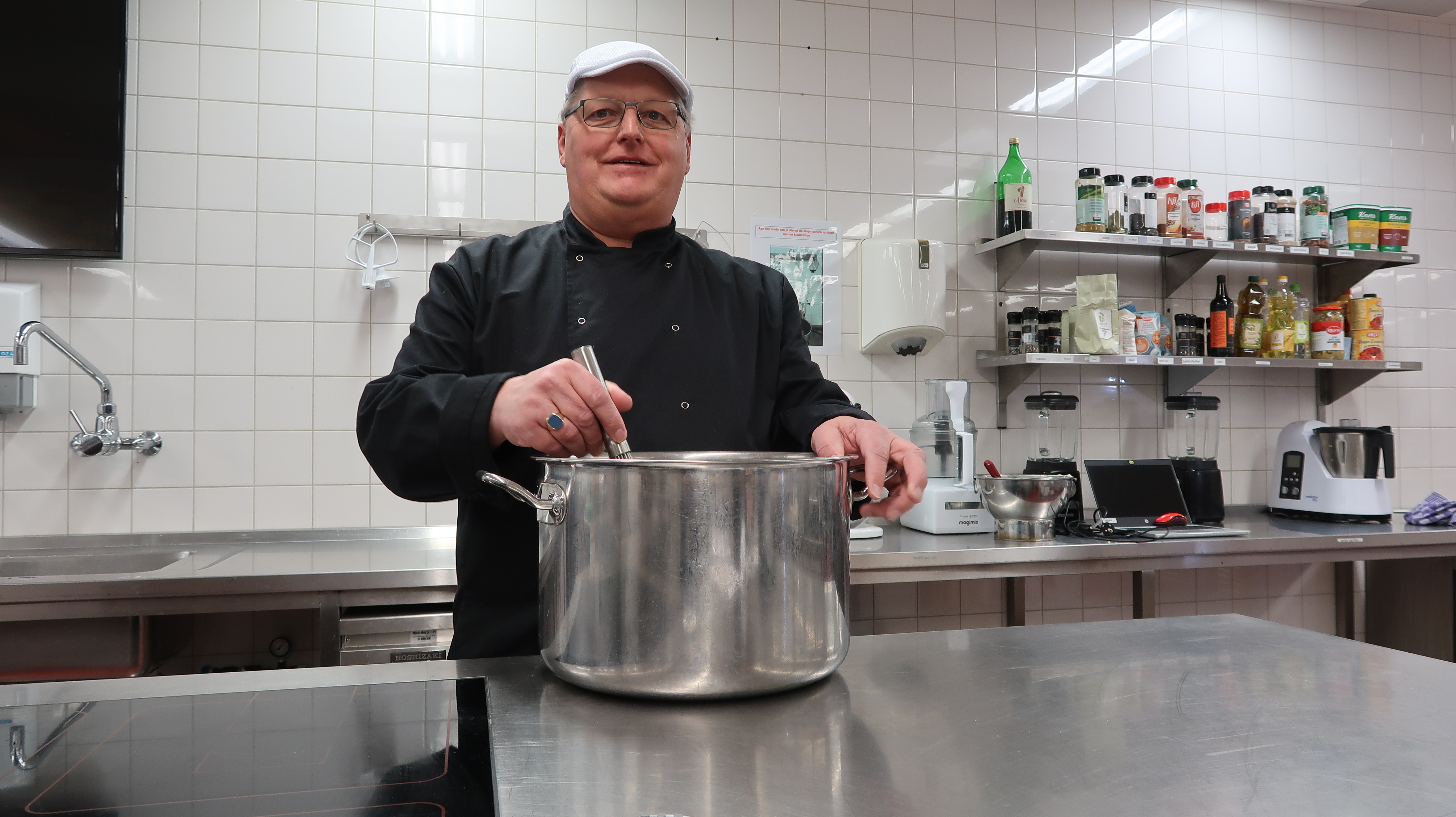 Cursist in een professionele keuken bereidt maaltijd in grote kookpan op het fornuis.