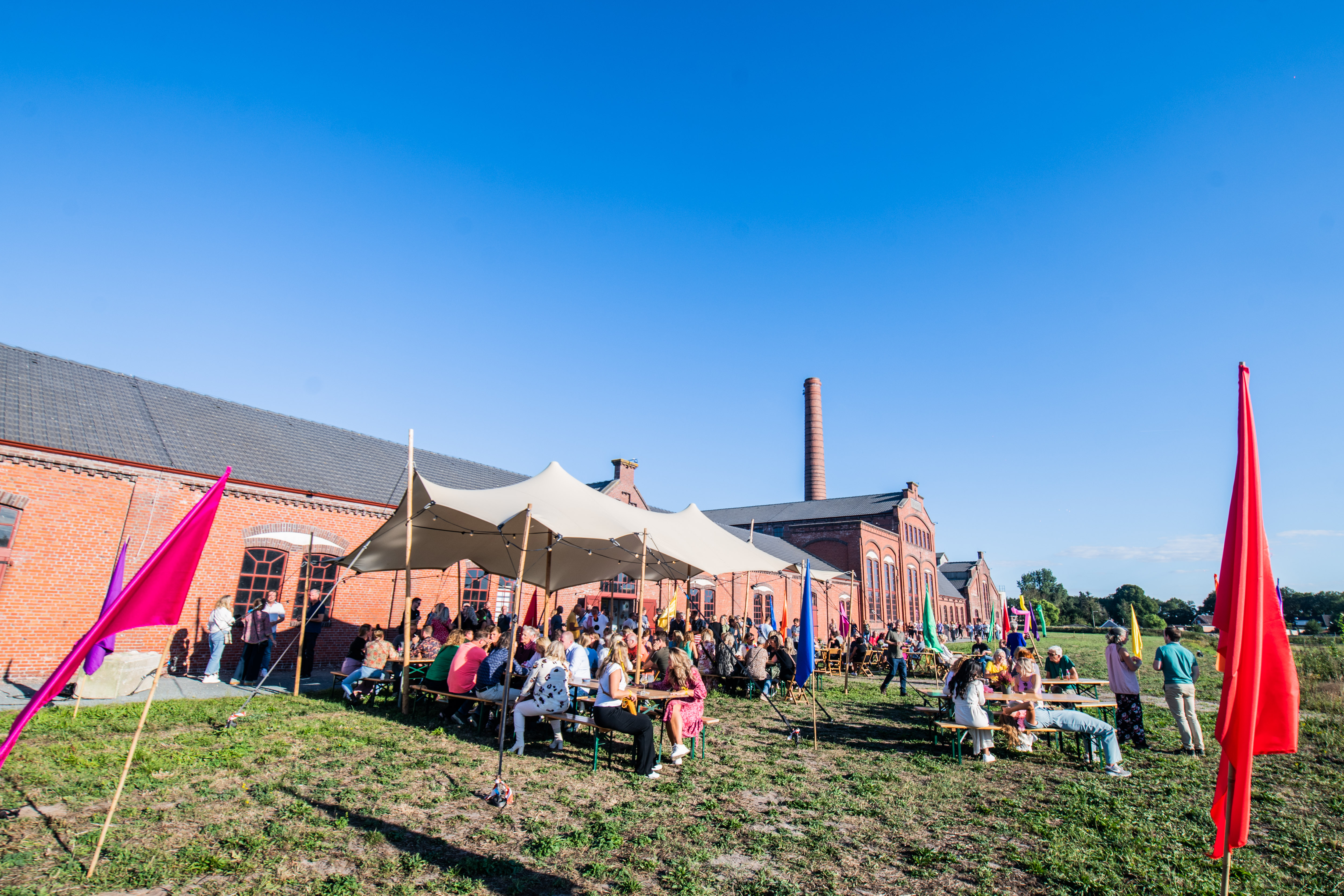 Groep studenten en bezoekers verzamelen zich buiten voor een jubileumevenement bij een industrieel gebouw.