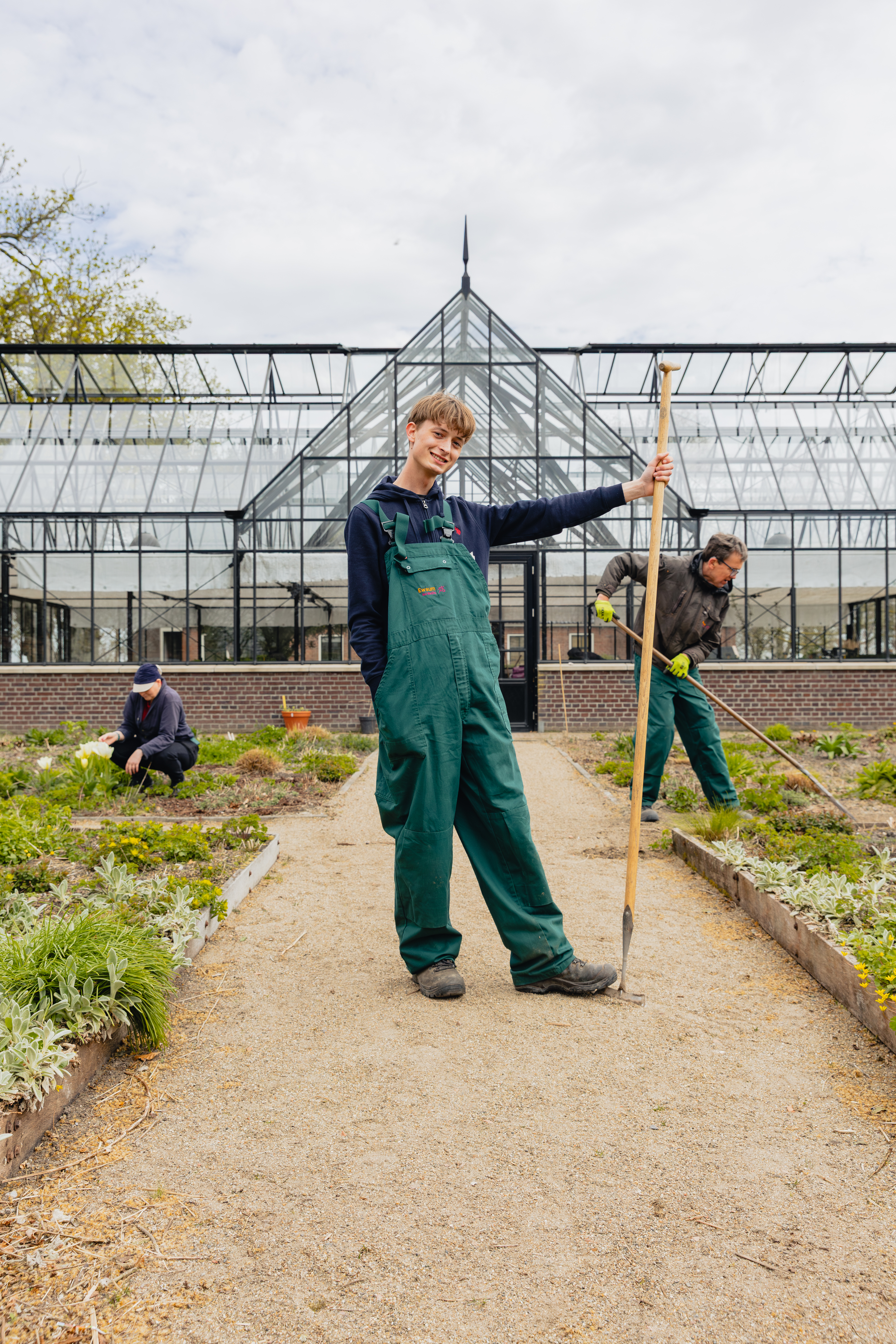 Leerling houdt een tuinhark vast in een tuin bij een kas, terwijl anderen planten verzorgen tijdens een stage bij WerkPro Op Ewsum.