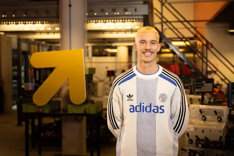 Student poseert in technische leeromgeving met machines en een opvallend gele pijl op de achtergrond