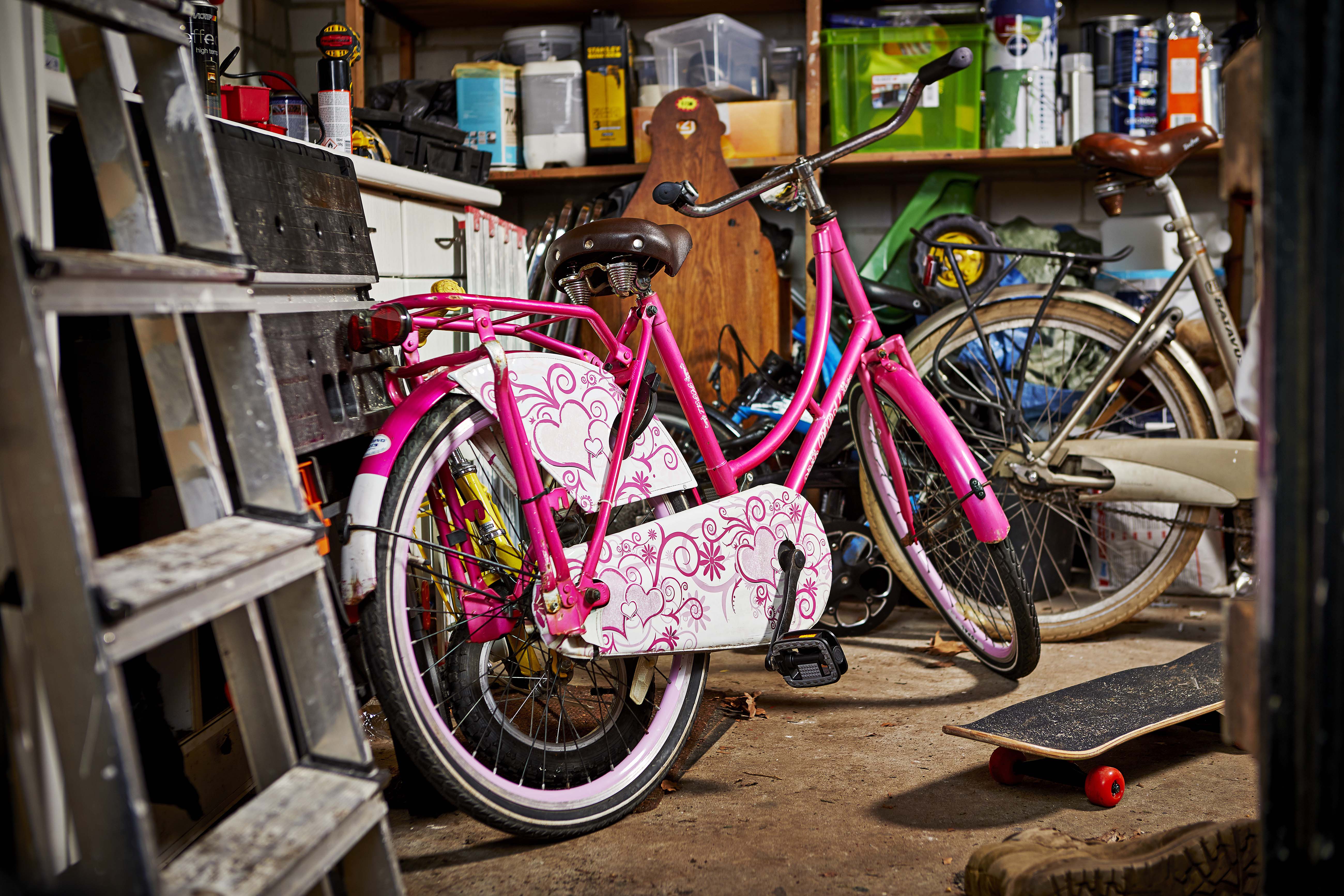 Roze kinderfiets met bloemmotief staat in een werkplaats tussen andere fietsen en een skateboard