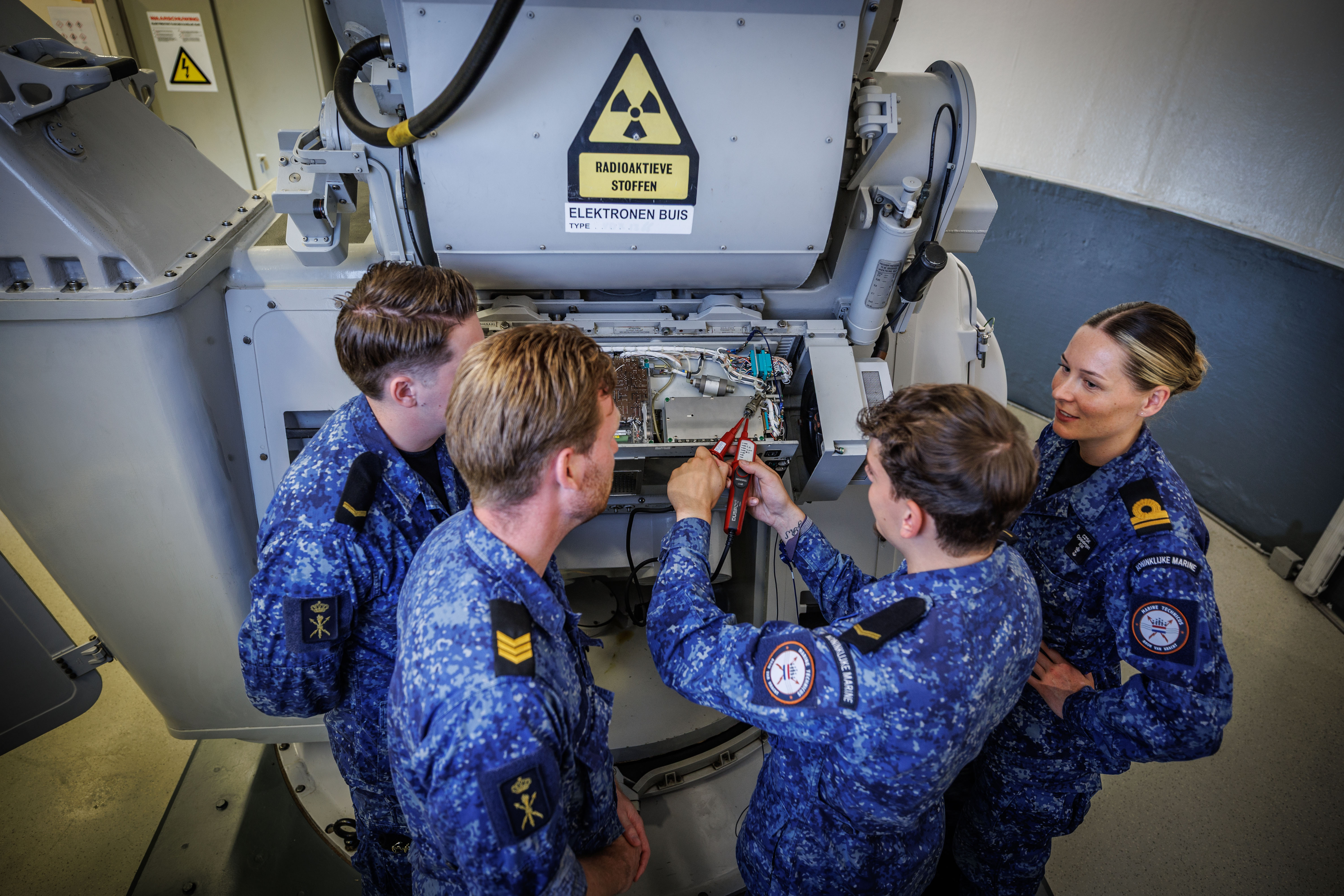 Vier deelnemers in technische uniformen werken samen aan het onderhoud van een elektronisch apparaat.