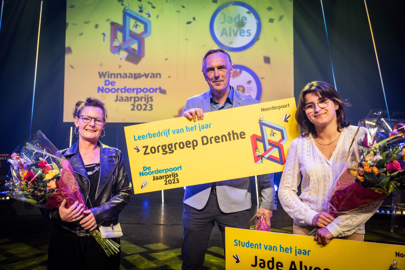 Drie personen tijdens uitreiking Noorderpoort Jaarprijs 2023 met borden en bloemen op het podium