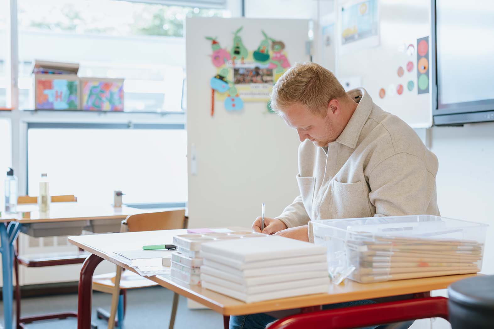 Onderwijsassistent werkt geconcentreerd aan schrijfopdracht in een lichte klasomgeving met educatief materiaal.