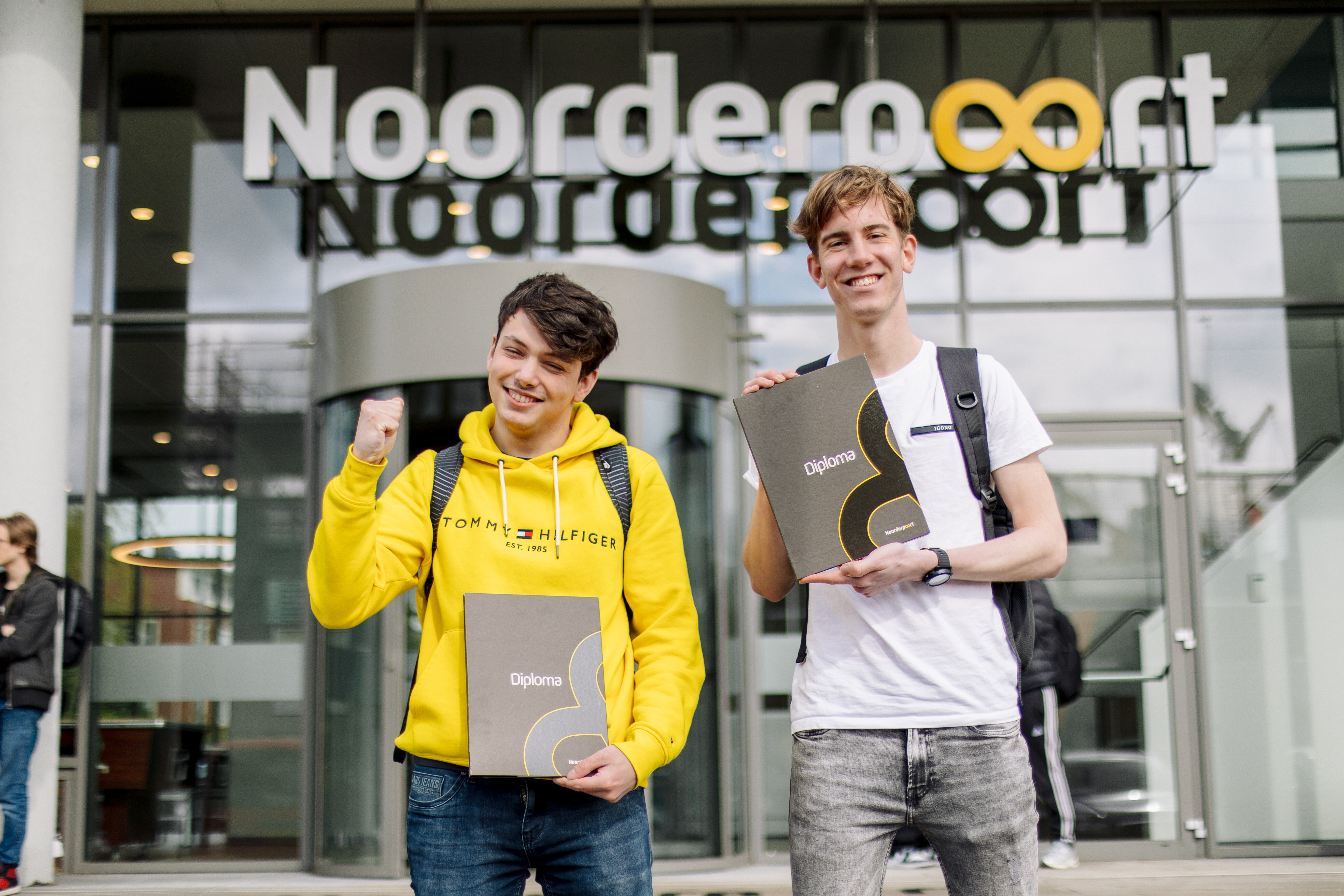 Twee studenten tonen trots hun diploma's voor het Noorderpoort-gebouw na het afleggen van hun examen