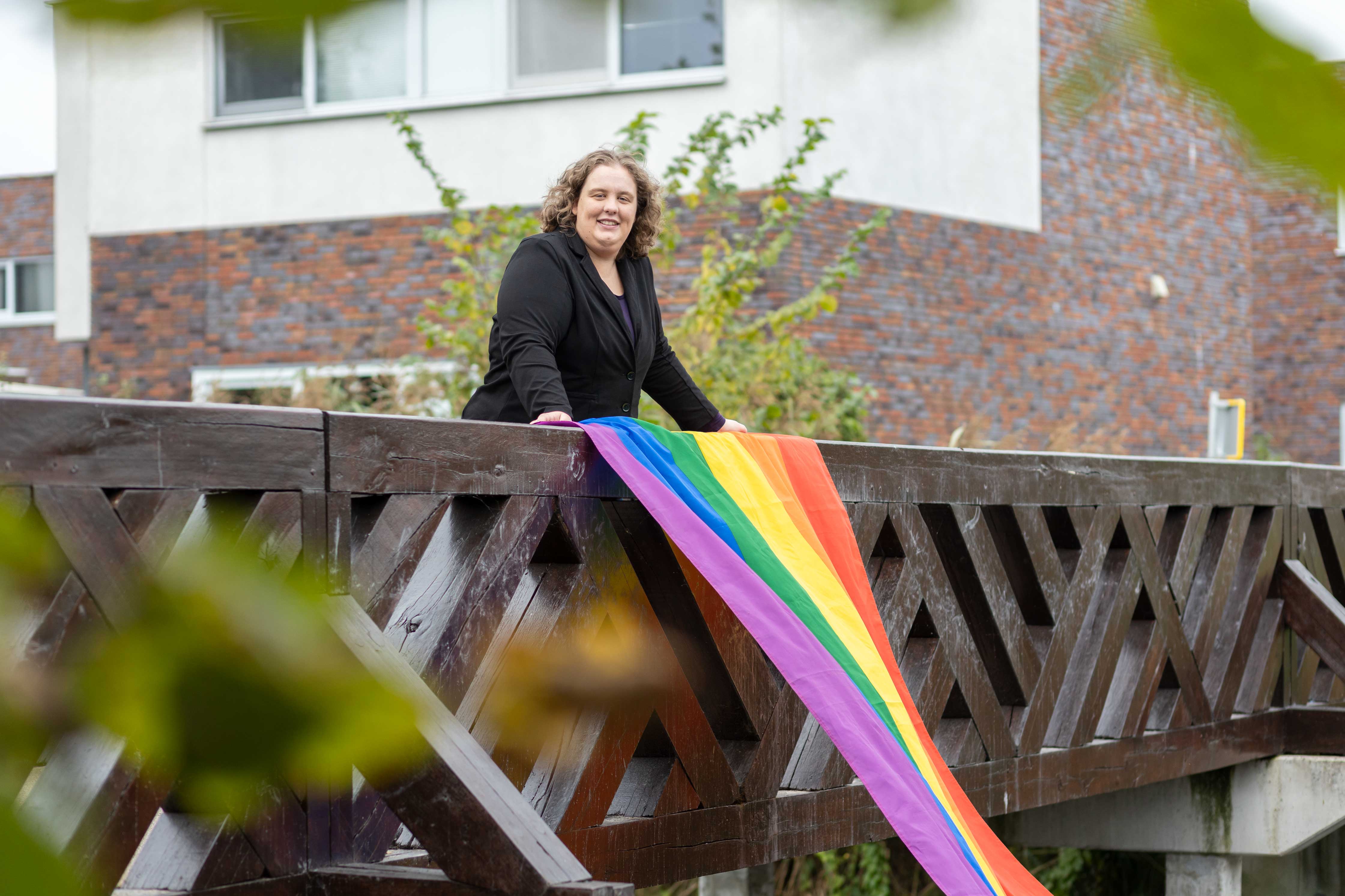 Persoon leunt op houten brug met regenboogvlag, symbool voor acceptatie en diversiteit.