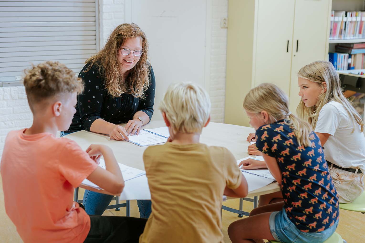 Onderwijsassistent begeleidt groep leerlingen bij een groepsopdracht aan tafel in een klaslokaal.