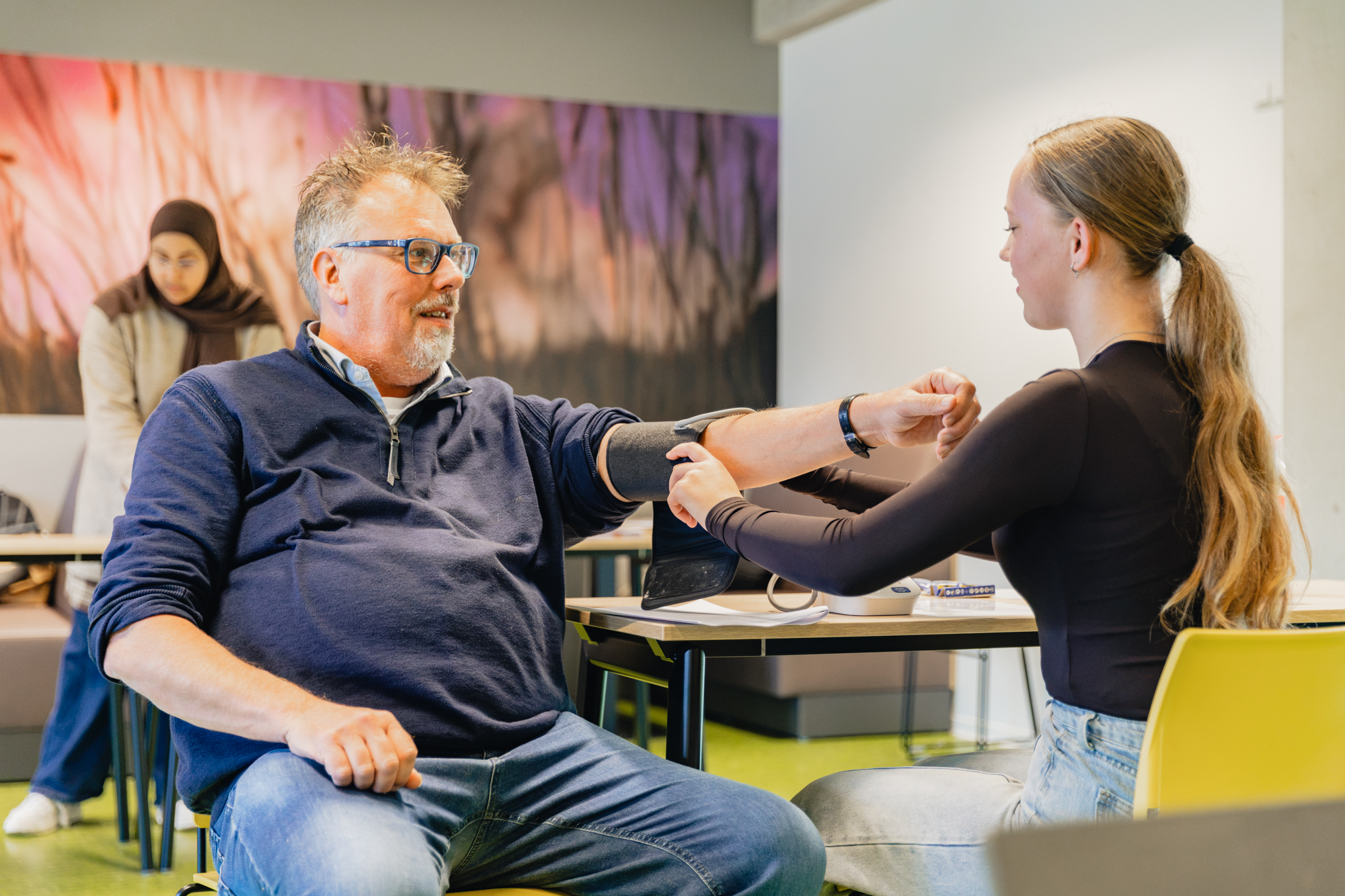 Student organiseert een fittest en meet de bloeddruk van een deelnemer van 55-plus in een klaslokaal