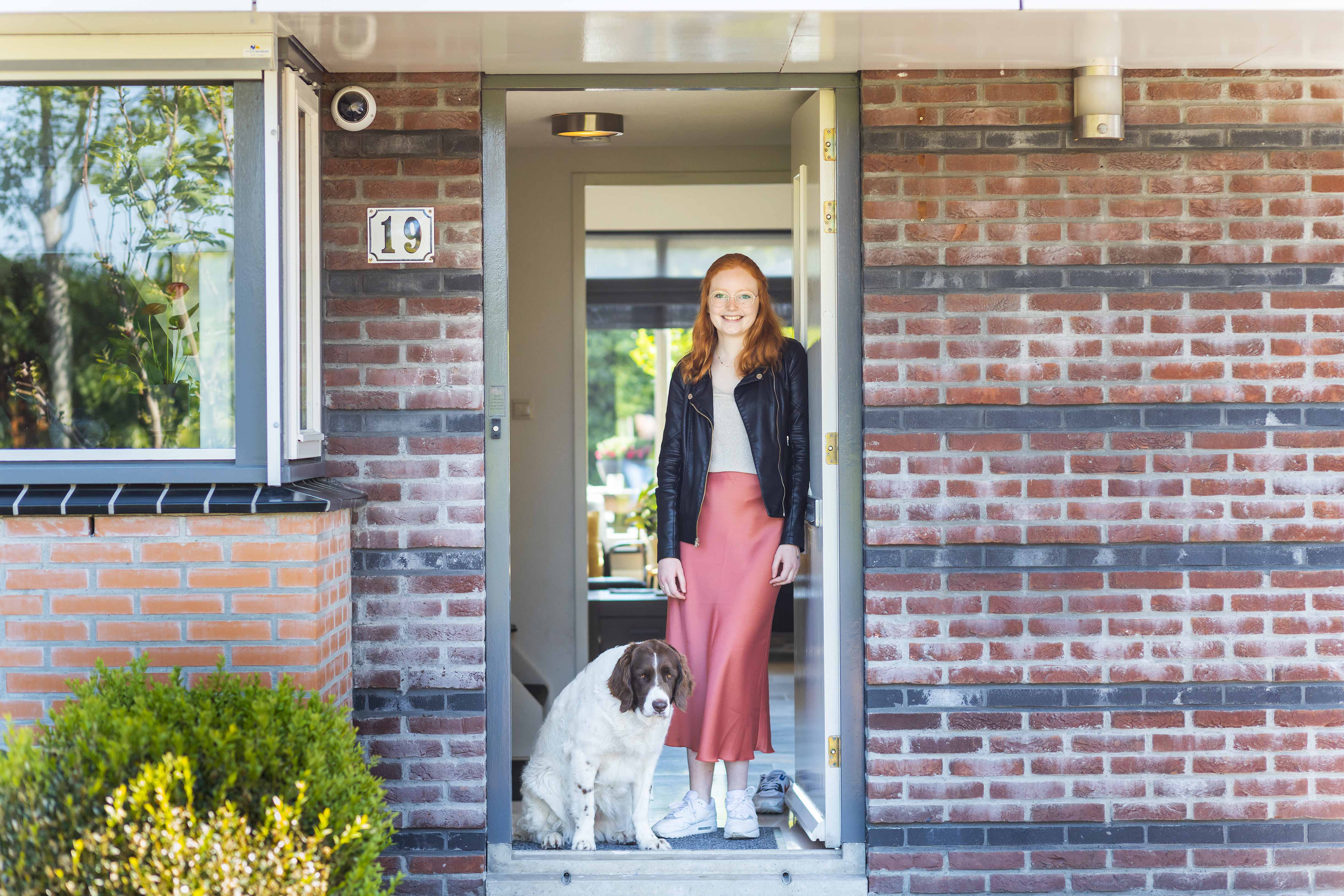 Student staat in de deuropening van een woning met een hond aan haar voeten, symbool voor succes en persoonlijke groei