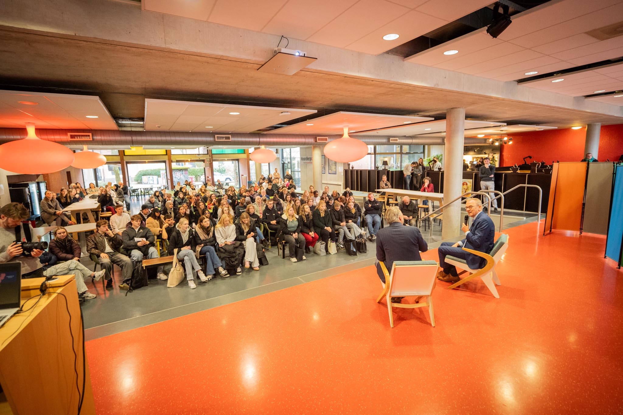 Gesprekssituatie met minister Bruins en een interviewer op het podium voor een toehoordersgroep studenten bij Noorderpoort