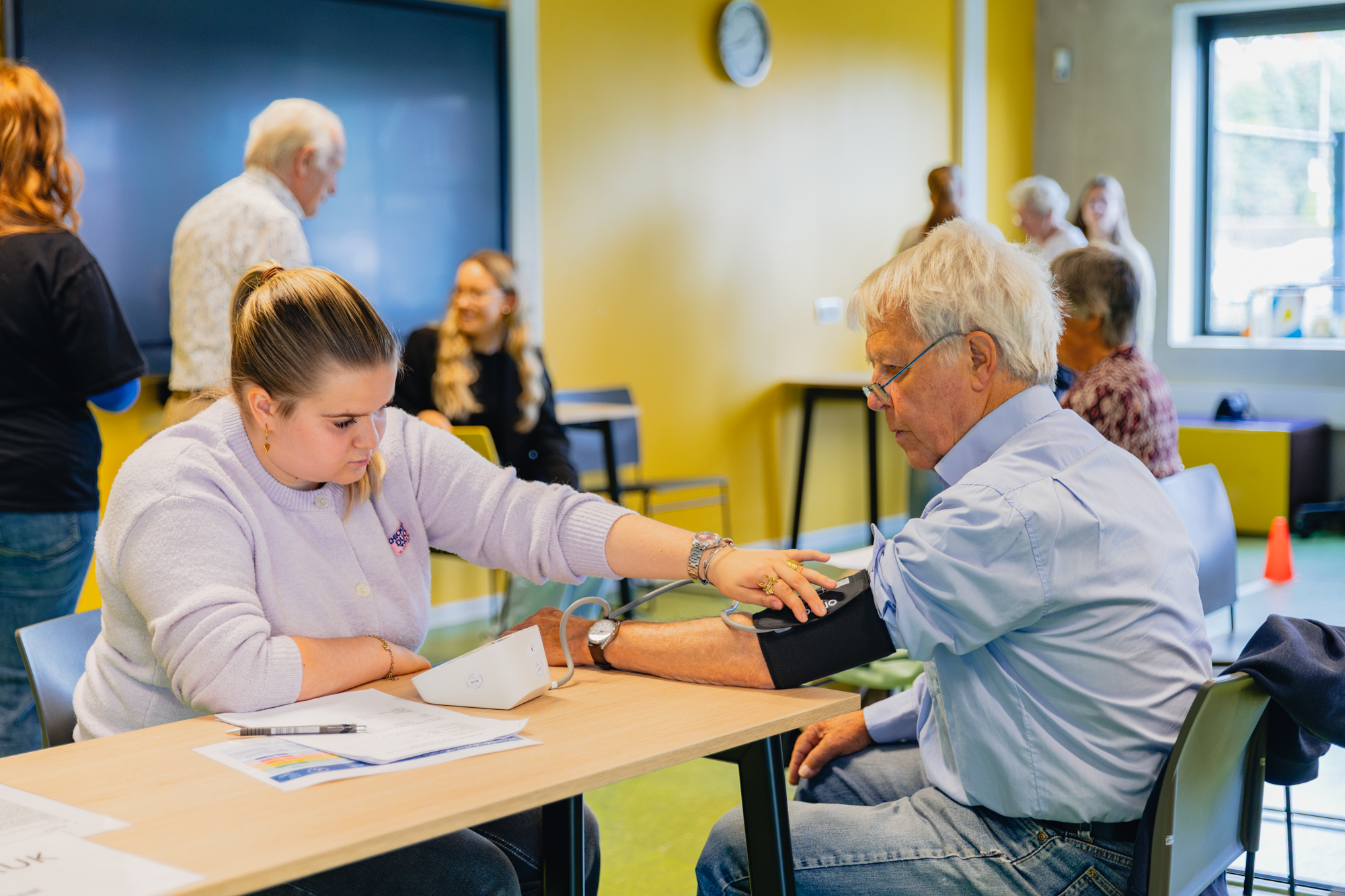 Student meet bloeddruk bij deelnemer tijdens fittest voor 55-plussers in een lesruimte