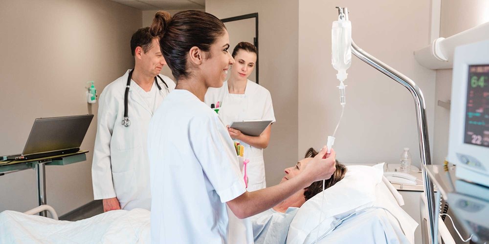 Verpleegkundige controleert infuus bij patiënt terwijl collega’s de gezondheidstoestand bespreken