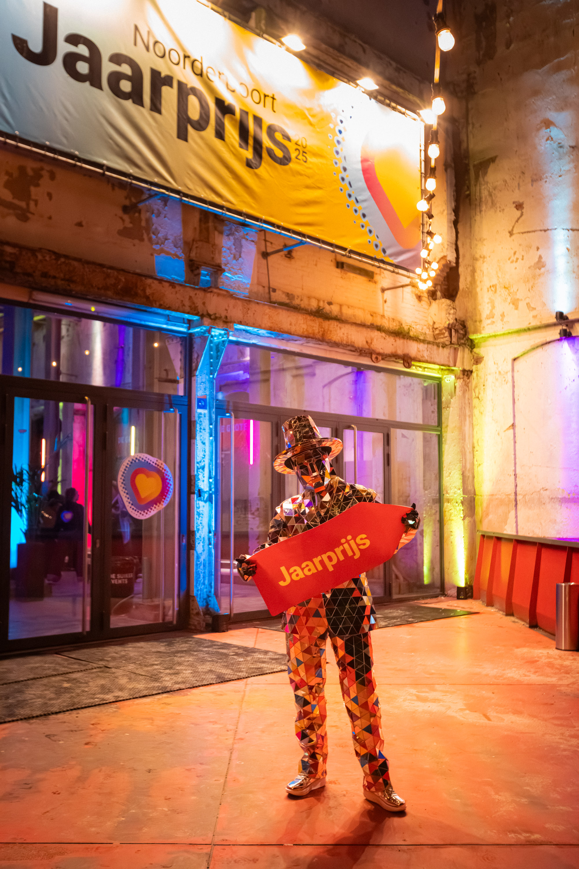 Persoon in kleurrijk kostuum houdt bord met tekst 'Jaarprijs' vast tijdens evenement van Noorderpoort
