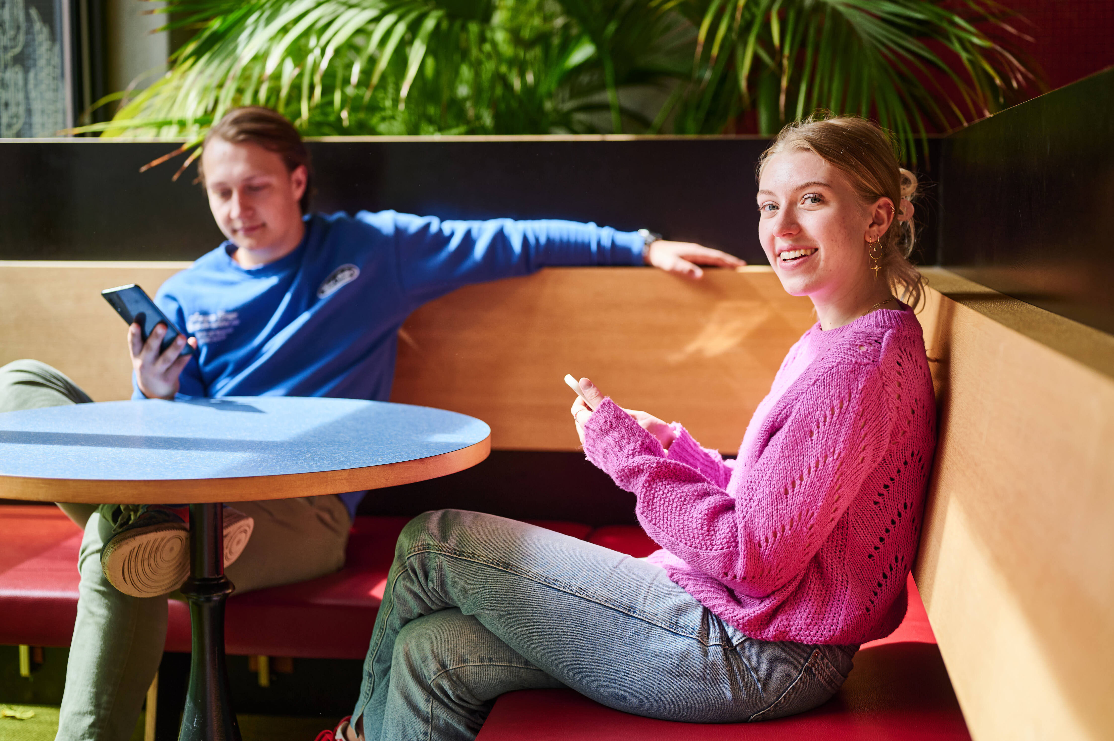Studenten zitten ontspannen in een zithoek, één bekijkt een mobiel apparaat, de ander is in gesprek.