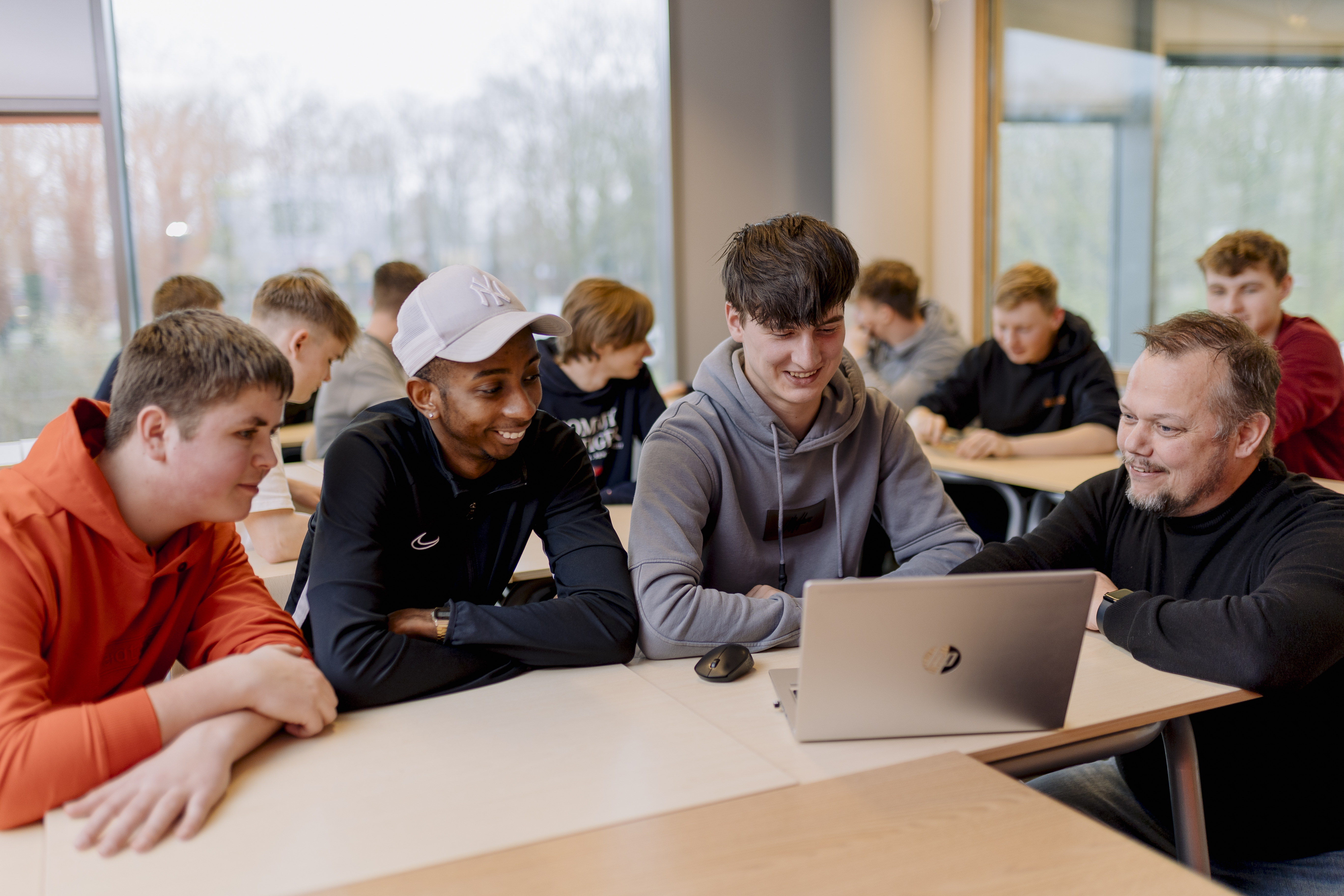 Docent begeleidt groep studenten tijdens interactieve les met laptop aan tafel in klaslokaal