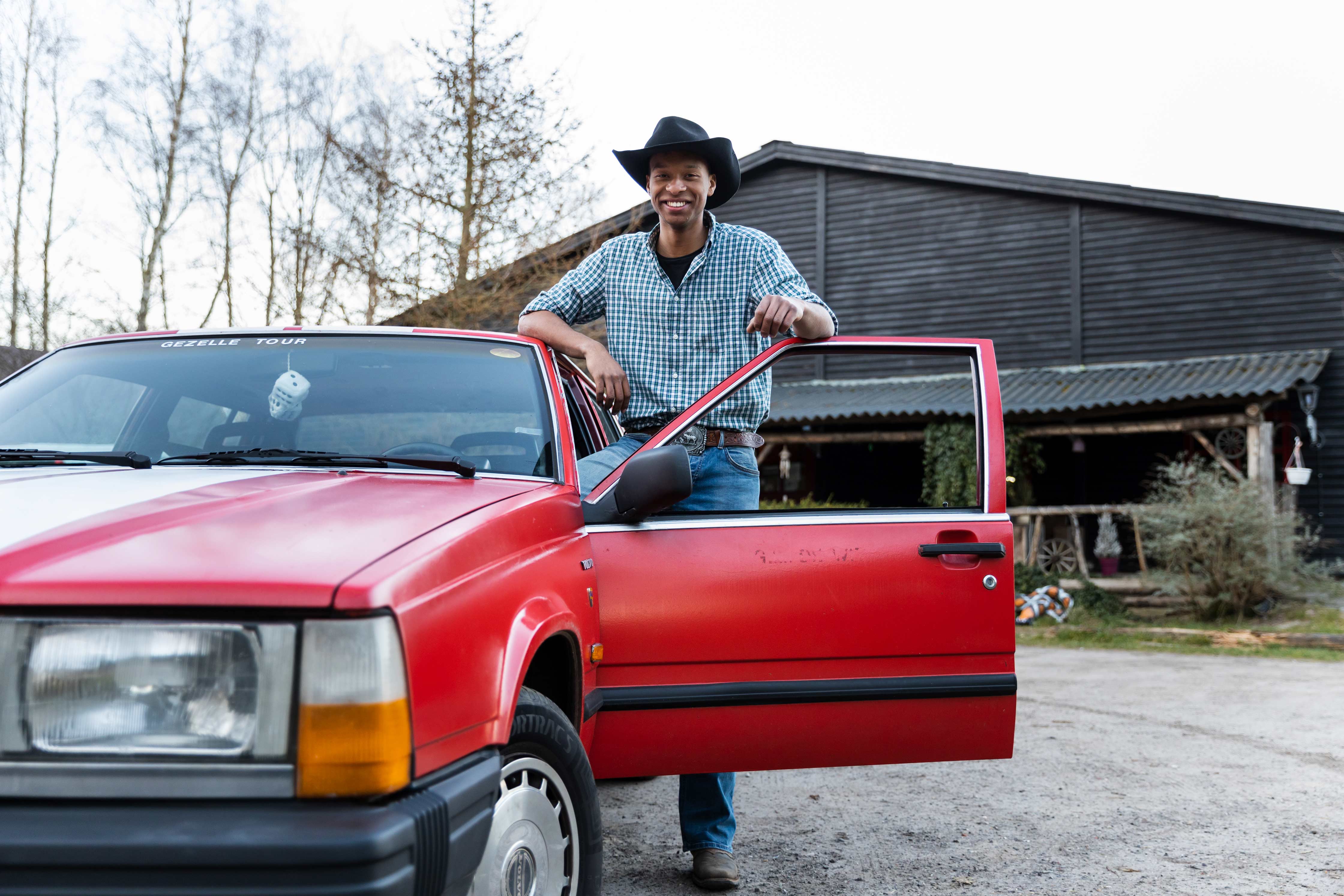 Student die naast een rode auto staat en een cowboyhoed draagt, passend bij het thema bijzondere combinatie.